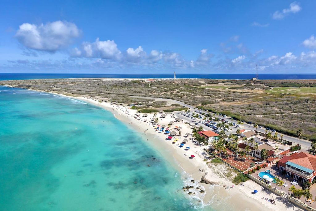 This aerial view shows a stunning stretch of coastline in Aruba, with the crystal clear waters of the Caribbean Sea lapping at the white sandy beaches. Arashi Beach is in the picture's foreground, a beautiful crescent-shaped beach with calm, turquoise waters. In the distance, rising from a rocky promontory is the iconic California Lighthouse.